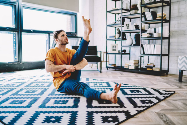 Young man stretching leg and looking up doing sportive exercises during morning workout in modern apartment with home interior,Hipster guy develop flexibility and doing yoga on comfortable carpet - Фото, изображение