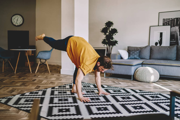 Young man holding leg up during morning yoga exercises on cozy carpet at home apartment with stylish interior.Hipster guy lead healthy lifestyle doing stretching training in stylish flat - Фото, изображение