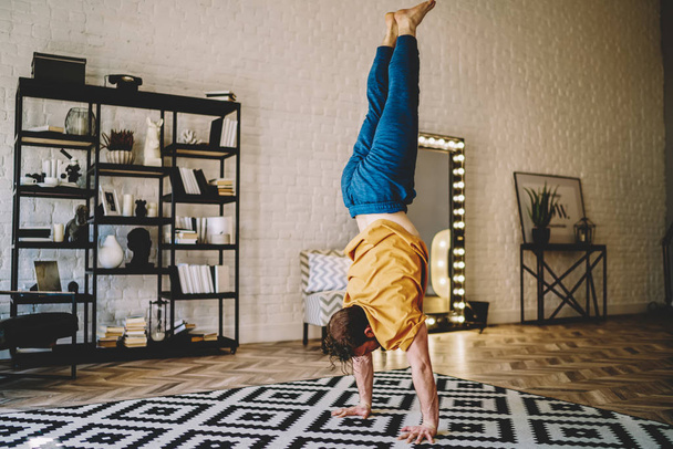 Motivated young man standing on his hands doing morning exercises during yoga at home interior.Male lover of healthy lifestyle develop strength of hands practicing active training in apartment - Фото, зображення
