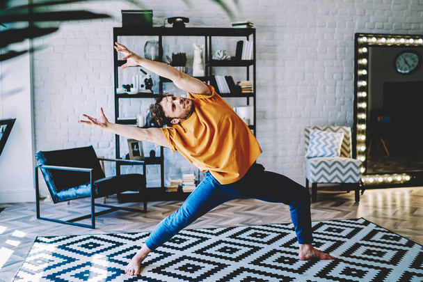 Motivated young man bends and stretching his hands to the side during morning exercises at home interior.Hipster guy doing yoga and lends healthy lifestyle standing on carpet in apartment - Fotó, kép