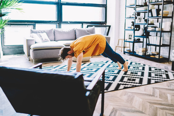 Motivated young man lover of yoga engaged in sport and leads healthy lifestyle at home apartment.Hipster guy practicing sportive poses on carpet to support body shape and vitality in comfortable flat - Photo, image