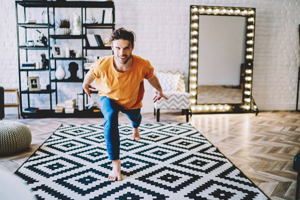 Portrait of positive young man smiling at camera while practicing different poses during yoga training at home interior.Motivated hipster guy warming up before meditation in modern apartment - 写真・画像