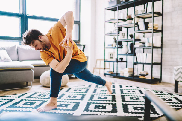 Motivated flexible young man in sportive wear engaged yoga training on stylish carpet in modern apartment in morning free time.Male lover of meditation standing in sport pose during workout - Foto, immagini
