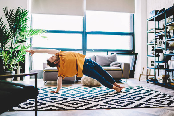 Experienced male lover of yoga in sportive wear stretching hand standing in yoga pose during morning training at home interior.Professional sportive man practising meditation in modern apartment - Фото, изображение