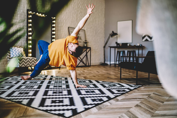 Professional male lover of yoga practising poses on stylish carpet in modern apartment during morning workout.Experienced young man doing sport exercises to support healthy lifestyle - Foto, immagini