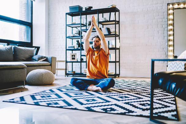 Calm young bearded man sitting in lotus pose on cozy carpet and holding hands in namaste above head during meditation at home apartment.Hipster guy with closed eyes doing yoga exercises - Zdjęcie, obraz