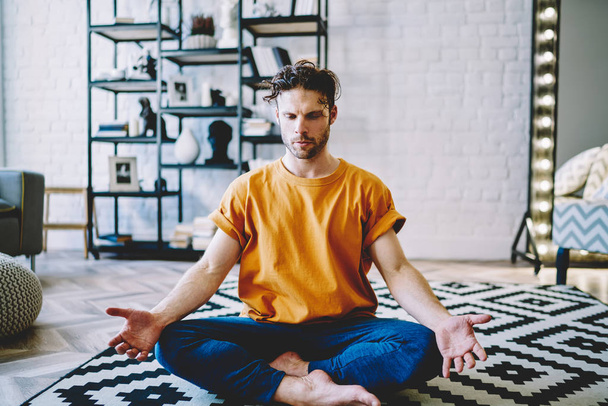 Concentrated young bearded man in active wear sitting in lotus pose during morning training on comfortable carpet in modern apartment.Calm hipster guy engaged in yoga at home interior - Photo, image