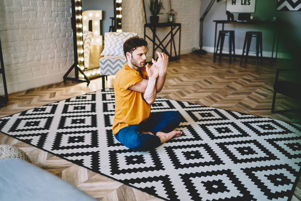 Young man stretching hands during asana pose sitting in lotus on comfortable carpet in modern apartment.Hipster guy 20s old practicing meditation during morning training at home interior - Foto, Bild