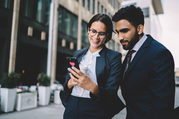 Successful cheerful female entrepreneur showing text on financial website with funny joke about management to spanish male partner standing near, concept of executive employees in business district - Foto, Imagen