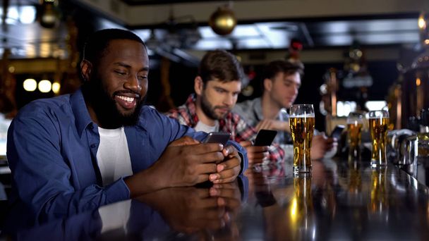 Smiling afro-american male sitting in pub looking through dating sites addiction - Foto, afbeelding