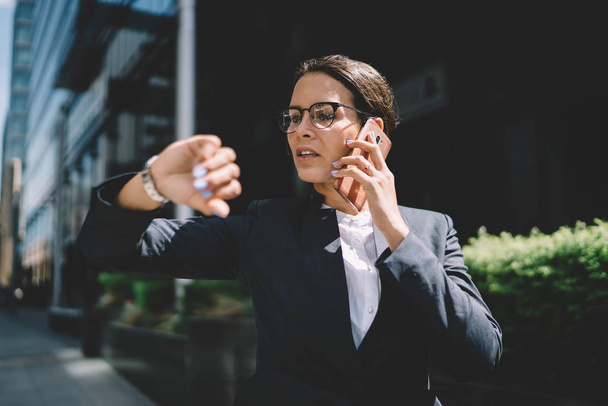 Udana właścicielka firmy, dzwoniąca do dyrektora finansowego, by omówić czas spotkania sprawdzona na noszonym zegarku, poważna kobieta w okularach korzystająca z połączenia roamingowego w telefonie komórkowym - Zdjęcie, obraz