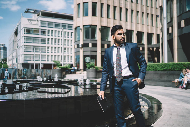 Successful male economist dressed in formal suit walking near modern fountain and thinking about upcoming interview meeting, pensive spanish man strolling on financial district with folder in hand - Foto, imagen