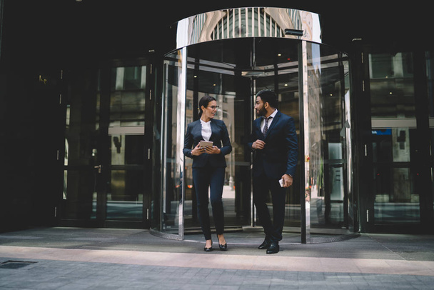 Positive male and female coworkers going out from office building for spending break together enjoying friendship togetherness, successful economists in stylish suits discussing cooperation - Foto, imagen