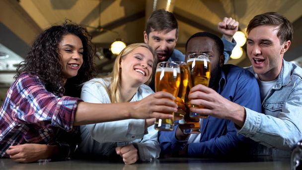 Cheerful friends clinking beer glasses, celebrating team victory in bar, leisure - Foto, Imagen