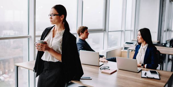 Confident female boss in formal outfit thoughtfully looking away holding cup while her male and female employee having friendly conversation at desktops during coffee break in loft office interior - Foto, immagini
