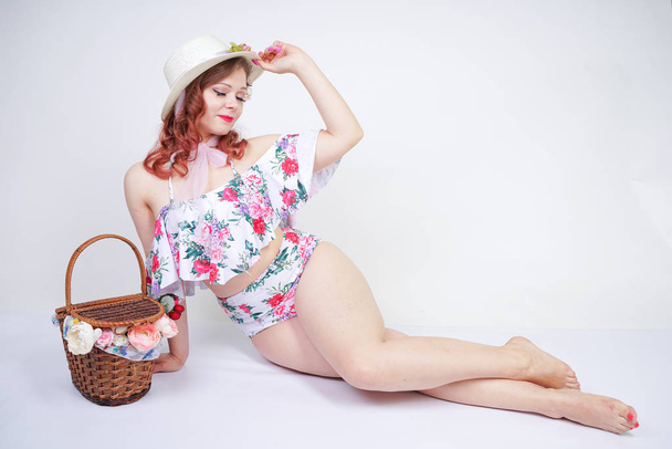 beautiful young pin up caucasian girl in romantic fashionable straw hat, vintage swimsuit with flowers and retro wicker basket posing on white background in Studio alone. pretty pinup woman relaxing. - 写真・画像