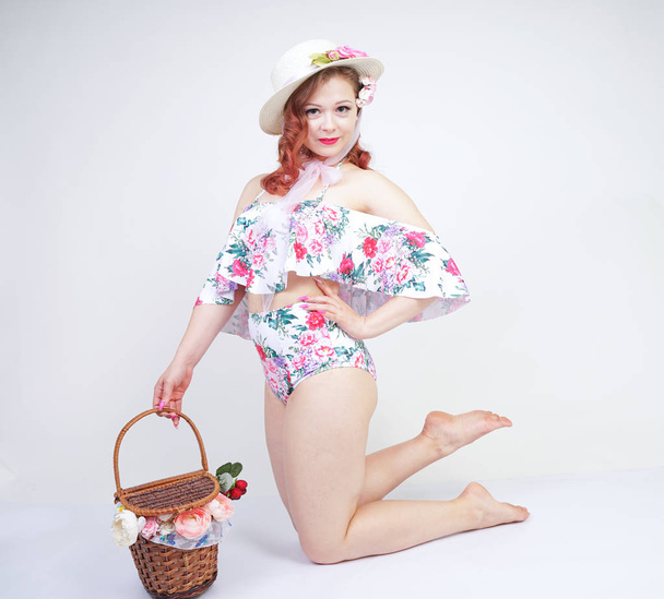 beautiful young pin up caucasian girl in romantic fashionable straw hat, vintage swimsuit with flowers and retro wicker basket posing on white background in Studio alone. pretty pinup woman relaxing. - Foto, Imagen