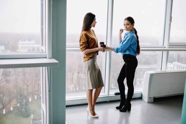 Full length portrait of female colleagues standing in loft interior office communicating, attractive caucasian businesswomen in trendy clothes having conversation about work during coffee break - Foto, immagini