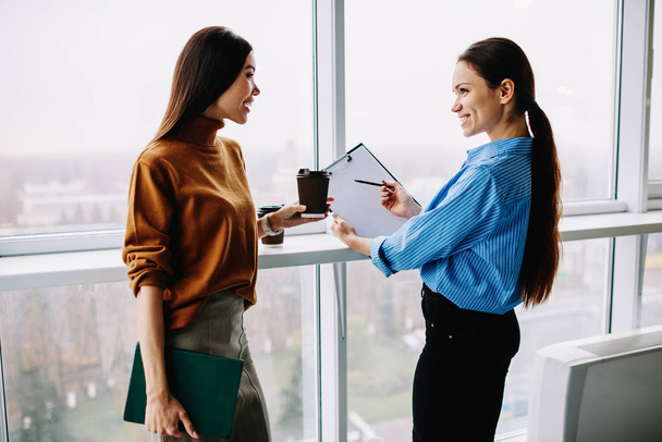 Cheerful female employee suggesting her colleague to sign papers having friendly conversation in office, women coworkers discussing planning for common project during free time of coffee break - Photo, Image