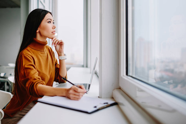 Thoughtful caucasian woman pondering  during planning project making notes in modern loft interior office, contemplative businesswoman looking at window thinking about future of finance company - Photo, Image