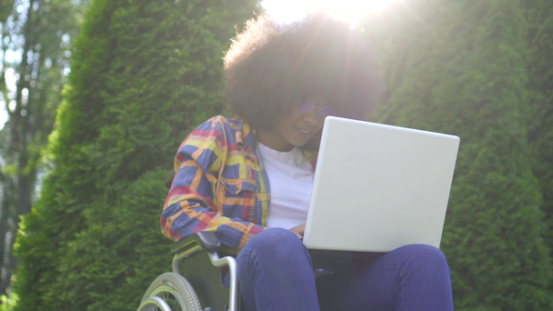 Mulher americana africana sorridente com um penteado afro desativado em uma cadeira de rodas usa um sunflare laptop no parque
 - Filmagem, Vídeo