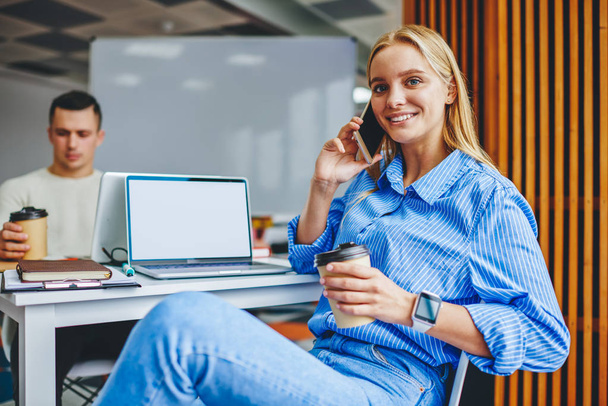 Portrait of charming positive young woman with coffee in hand calling on smartphone while smiling at camera.Cheerful female manager talking on cellular sitting at desktop with laptop with blank screen - Zdjęcie, obraz