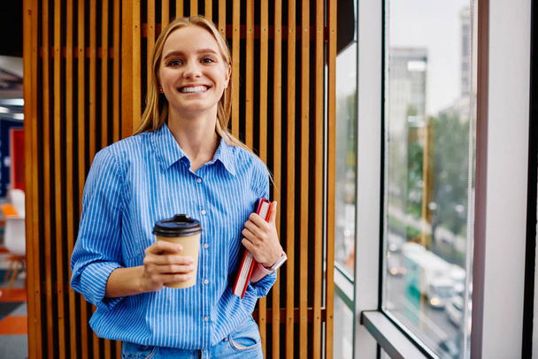 Portrait of positive hipster student with notebook smiling at camera.Cheerful young woman holding tasty coffee to go while standing in modern office interior.Happy female employee with cup of caffeine - Photo, Image