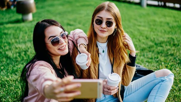 Brunette and blonde female bloggers posing for smartphone camera spending summer time in park, hipster girls friends enjoying coffee break together taking photos for share in social networks profile - Zdjęcie, obraz