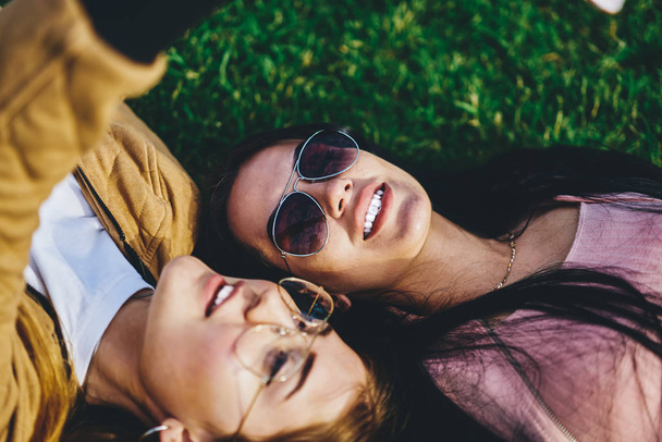 Close up photo of attractive brunette and blonde female friends lying on green grass in trendy eyewear and sunglasses, beautiful hipster girls recreating in park together during spring leisure - Fotoğraf, Görsel