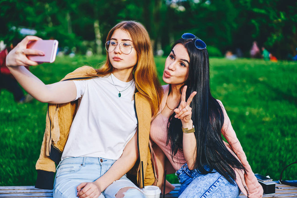 Attractive brunette and blonde female friends in casual stylish wear posing for selfie using mobile phone camera, hipster girls making picture for share in blog resting on free time in park - Foto, Bild