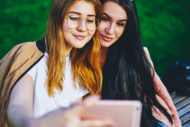 Attractive female best friends making selfie spending leisure together outdoors and using mobile phone camera, positive young women looking at smartphone taking photo via application in park - Foto, afbeelding