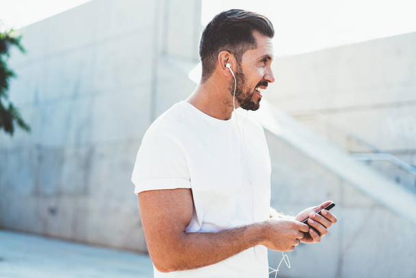 Side view of Caucasian man dressed in white t shirt with copy space area for brand label holding modern cellphone gadget in hand listening music playlist and looking away, concept of technology - Photo, Image