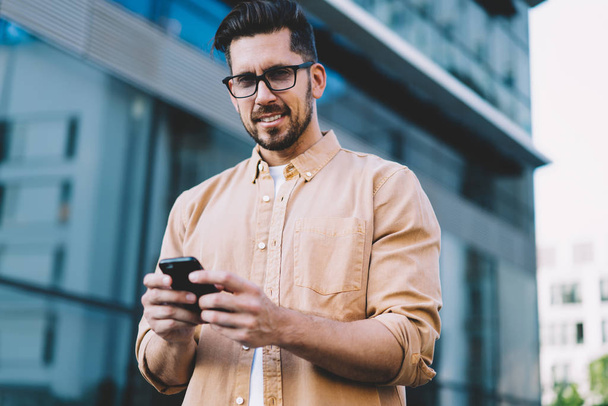 Portret przystojnego mężczyzny rasy kaukaskiej z nowoczesną technologią komunikacji stojącego na miejskim planie i pozującego, brodatego użytkownika smartfona w okularach optycznych do korekcji wzroku patrzącego w kamerę - Zdjęcie, obraz