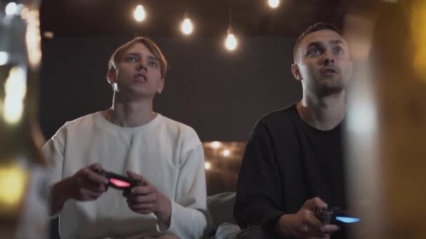 Two happy friends playing video game in the gaming room sitting on the sofa. Friends spending time together indoors. Video game and leisure concept. - Video, Çekim