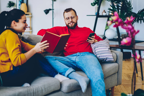 Young family of positive woman and man discussing new novel plot sitting on couch during common leisure, romantic couple spending free time on hobby and communication at comfortable apartmen - 写真・画像