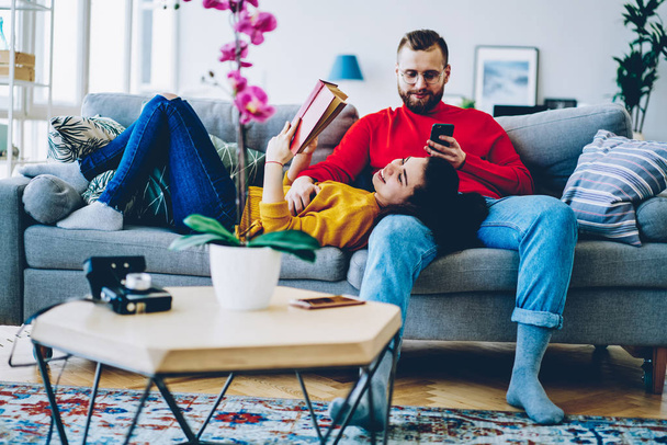 Young family resting on weekend at home together sending time on hobby , woman reading interesting book while her boyfriend using smartphone for checking mail in social networks on cozy couc - Foto, afbeelding