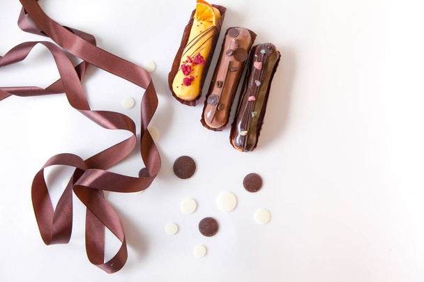 Un conjunto de tres eclairs con diferentes rellenos y diseño aislado en una superficie blanca decorada con gotas de chocolate y cinta de seda marrón
 - Foto, imagen