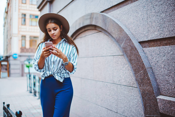 Γυναίκα τουρίστρια με μοντέρνα ρούχα και καπέλο χρησιμοποιώντας εφαρμογή στο smartphone για πλοήγηση στην πόλη, hipster κορίτσι ντυμένο με ρούχα μόδας έλεγχο μήνυμα μέσω κινητού τηλεφώνου και 4g σύνδεση σε εξωτερικούς χώρους - Φωτογραφία, εικόνα