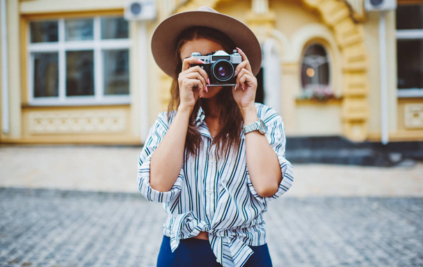 Young woman in trendy hat and apparel using vintage camera for taking picture spending time on street, young professional female photographer making image of urban settings on free time outdoors - Photo, Image