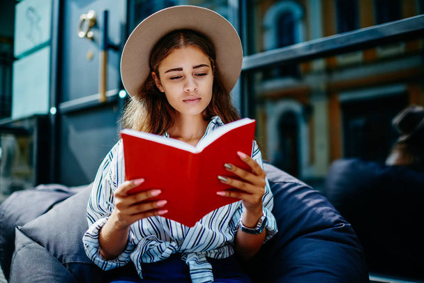 Pensive teen hipster girl in stylish hat sitting with interesting book outdoors satisfied with leisure time, beautiful young woman concentrated on interesting novel bestseller during free time - Photo, Image