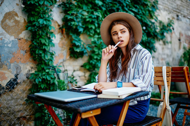 Pensive hipster girl in trendy hat pondering while creating article for blog holding pen in mouth sitting outdoors in cafe, brunette caucasian woman author writing in notebook making to do list - Foto, Bild
