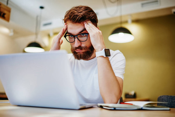 Sad male freelancer in eyewear for vision protection working overtime on laptop computer before deadlines, tired hipster guy feeling headache because of problems with online project on netbook - Φωτογραφία, εικόνα