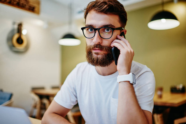 Portrait of serious caucasian bearded guy calling to banking service via mobile phone sitting indoors, young male hipster in eyewear for vision correction looking at camera having telephone talk - Foto, immagini