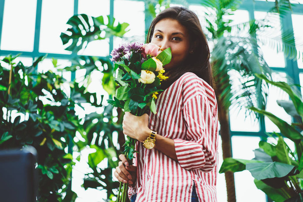 Attractive asian floral professional looking at camera and holding bouquet of tender asters and roses, young saleswoman posing in glasshouse with flowers in hands, concept of blooming composition - Foto, Imagen