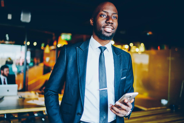 Successful African American entrepreneur dressed in suit smiling while looking straight. Prosperous male proud ceo i standing in modern office interior with smartphone in hands, Planning and dreaming - Foto, afbeelding