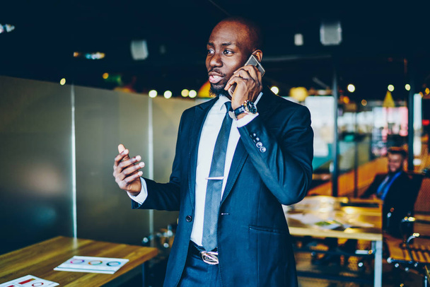 Dark skinned male entrepreneur dressed in suit discussing business plan during mobile conversation on smartphone device.African American proud ceo calling on cellular device while working in office - Foto, Imagen