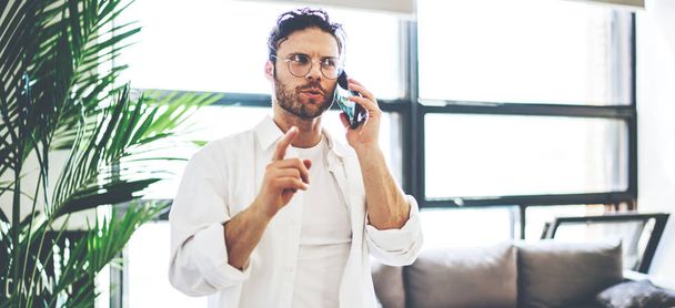 Young caucasian man in spectacles having mobile phone conversation standing at home interior, handsome hipster guy making booking via smartphone call choosing food delivery for apartment - Zdjęcie, obraz