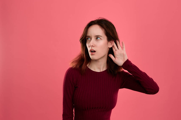 Surprised young lady standing isolated over dark pink background with hands raised close to ear as if is listening to some rumors. - Foto, Imagem