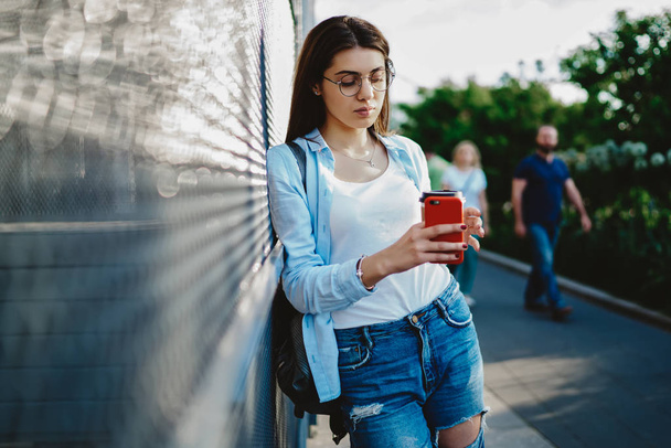 Caucasian young woman standing on street in urban setting and updating profile in social networks for online chat on modern smartphone device using high speed public 4G internet connection - Photo, image