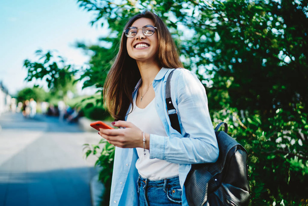 Positive hipster girl with backpack having fun outdoors in green park.Cheerful young caucasian woman holding digital mobile phone standing in urban setting enjoying good weather and great mood - Foto, imagen